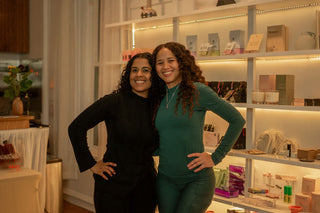 Two women standing side by side in front of shelves filled with various items.