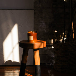 A candle on a wooden stool in front of a window, casting a warm glow in a cozy room.